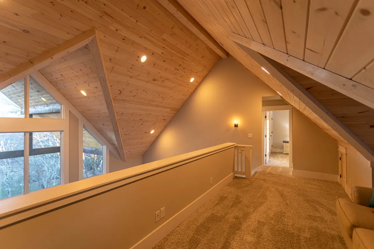 A tranquil, loft-like space under angled wooden ceilings, featuring soft carpeting and large windows that flood the area with morning sunlight. The open doorway hints at the versatile use of this area for relaxation, reading, or socializing.
