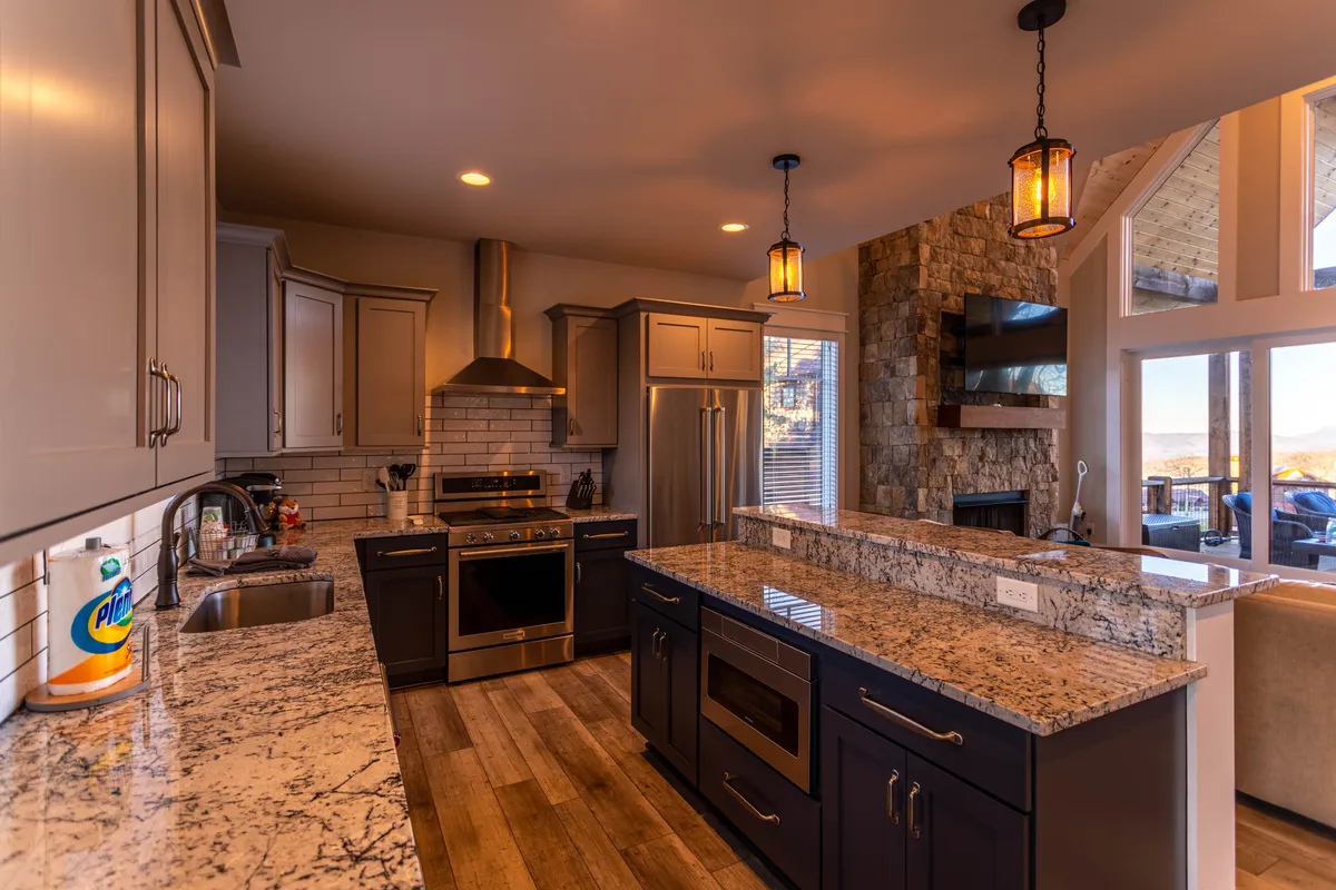 A warm, open-plan kitchen with expansive granite countertops, modern stainless steel appliances, and large windows that let in natural light. The adjacent stone fireplace and visible dining area hint at a seamless transition between cooking, dining, and entertaining, reinforcing the communal aspect of shared meals.