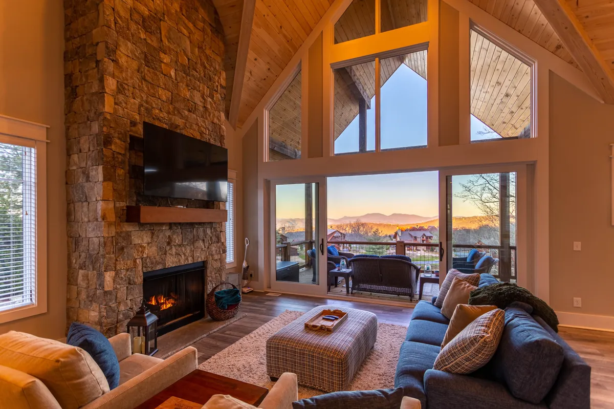 The image showcases a warm and inviting living room featuring a striking stone fireplace, expansive windows that frame stunning mountain sunsets, and a plush seating area perfect for relaxing or gathering with loved ones.