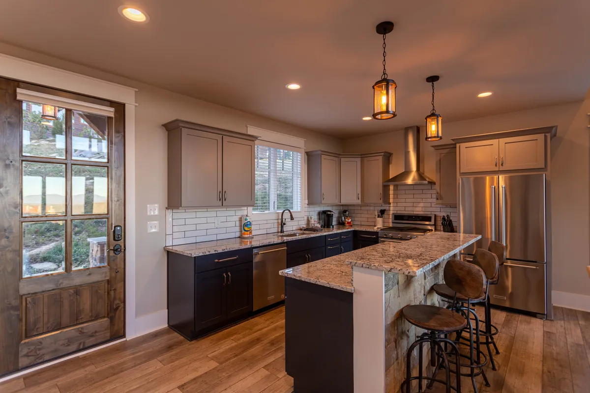A bright and welcoming kitchen scene featuring a spacious granite countertop, gleaming stainless steel appliances, and a well-appointed kitchen island. The wooden door and inviting layout suggest a space perfect for both meal preparation and entertaining, with subtle details like coffee mugs and cookware hinting at a fully stocked utensil set.
