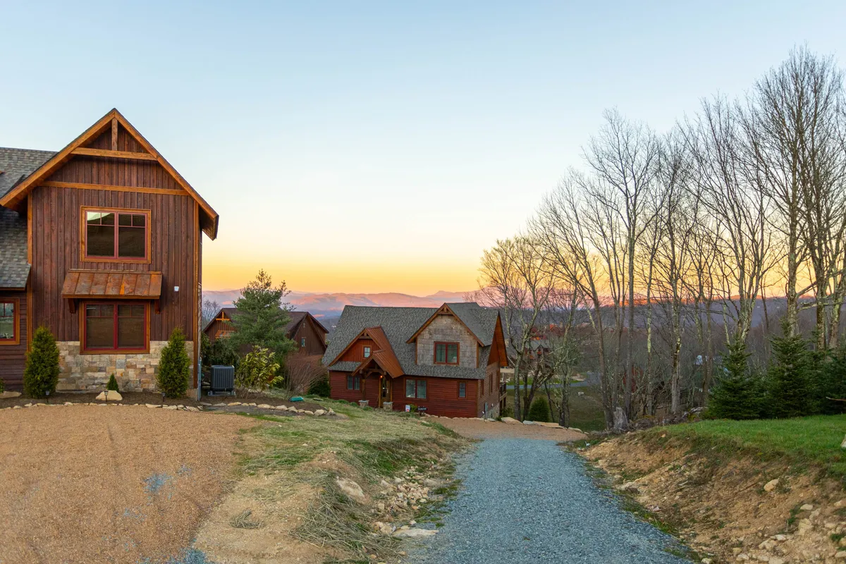 A serene view of the main house with a winding pathway and surrounding trees, all bathed in the warm glow of a sunset horizon. The setting suggests easy access to walking paths, communal spaces, and the natural amenities described in the section.