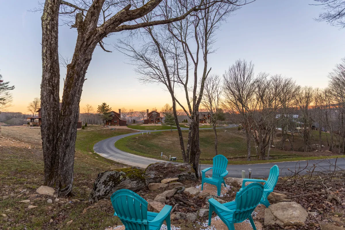 A cozy outdoor scene featuring teal Adirondack chairs positioned beneath tree branches, overlooking a winding road and distant cabins. The arrangement encourages conversation, stargazing, and evening gatherings around a fire.