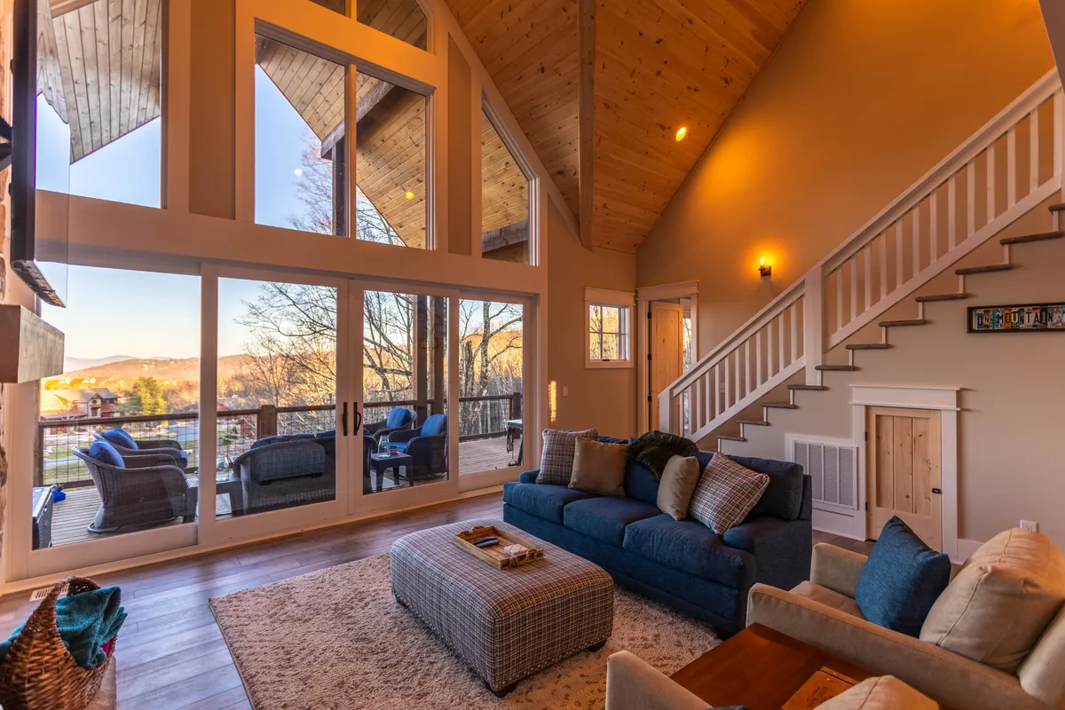 A cozy living room featuring a plush sofa positioned near expansive windows that offer a glimpse of the outdoors. The warm ambiance is perfect for relaxing with family or enjoying the view as the sun sets. The wooden staircase and decorative ottoman add to the inviting, comfortable vibe.