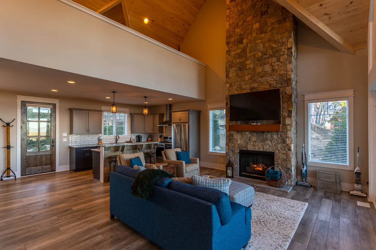 An inviting living area featuring a stone fireplace, ample windows revealing natural light and vistas, a large comfortable sofa, and a kitchen island visible in the background—suggesting effortless flow between living, dining, and kitchen spaces.