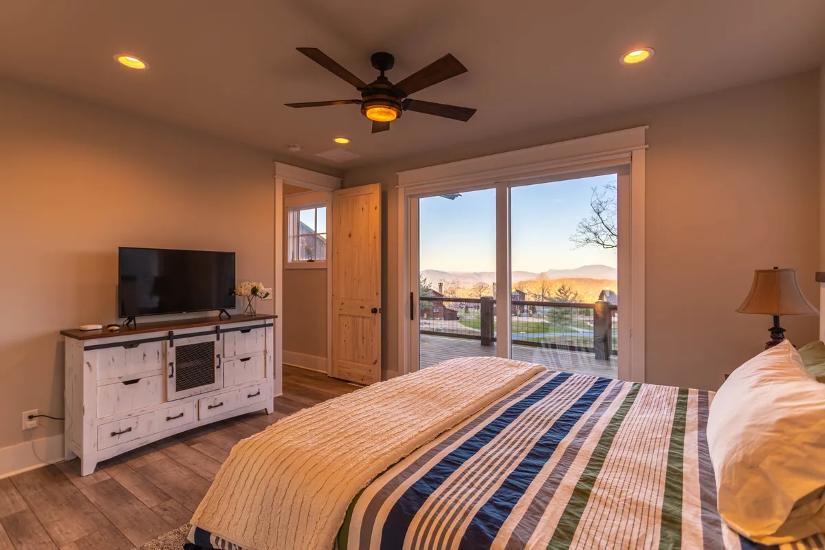 A cozy bedroom with a bed dressed in a striped blanket, a TV stand, and expansive windows offering scenic views. The setup invites relaxation, whether watching TV or simply taking in the mountain vistas from bed.