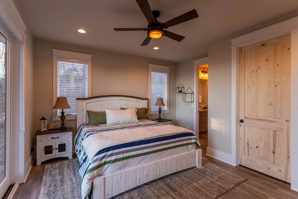 A calm and serene bedroom with a bed featuring a striped blanket, wooden bedside tables, and soft lighting from table lamps. Sunlight gently streams through the windows, emphasizing the inviting and restful atmosphere ideal for winding down after a day of adventure.